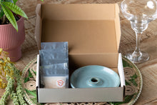 Load image into Gallery viewer, Open cardboard box containing a ceramic item and packets on a wooden surface with a plant and glassware in the background.