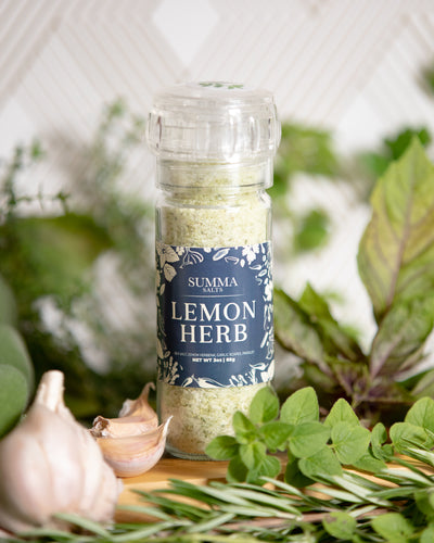 Summa Salts Lemon Herb Salt grinder surrounded by fresh herbs, lemon slices, and coarse sea salt, styled on a wooden board.
